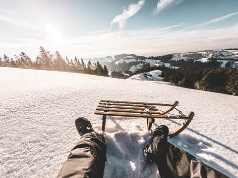 Pov view of young man looking the sunset on snow high mountains with vintage sledding - Legs view of travel influencer enjoying winter landscape - Vacation concept - Focus on wood luge