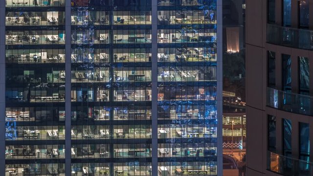Office building exterior during late evening with interior lights on and people working inside skyscraper night timelapse. Aerial close up view from above with many illuminated windows.