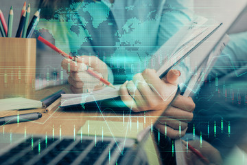 Fototapeta premium A woman hands writing information about stock market in notepad. Forex chart holograms in front. Concept of research. Multi exposure