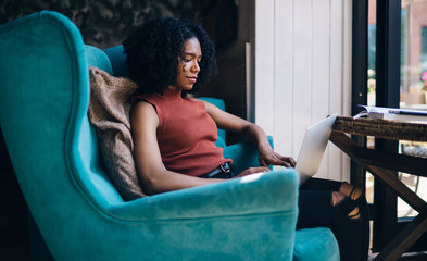 Positive female writer sitting in comfortable chair and using online application for typing content...