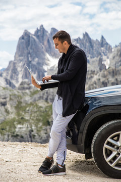 Young handsome man with laptop standing near his car and chek mail or working on beauty landscape of mountains. Work in vocation.