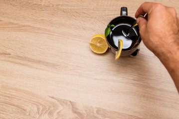 A mug of tea and lemons on the side with a hand steering spoon of sugar at a wooden table 