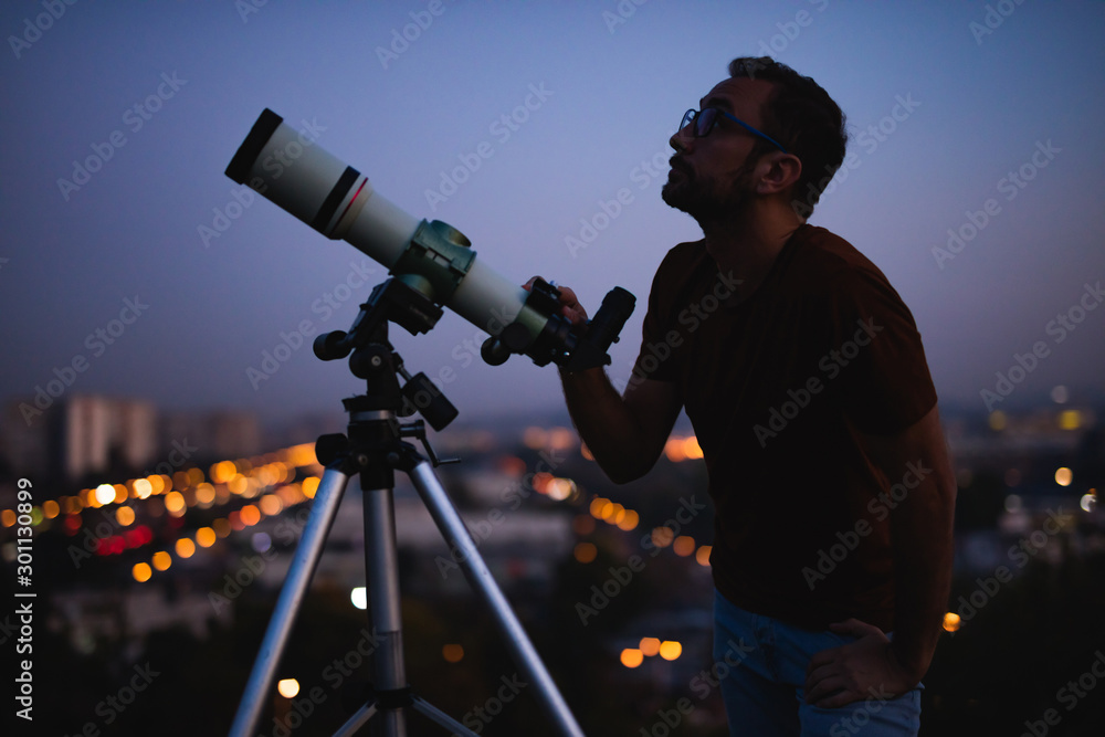 Wall mural astronomer with a telescope watching at the stars and moon with blurred city lights in the backgroun