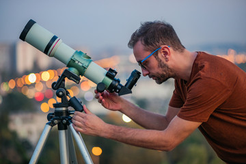 Astronomer with a telescope watching at the stars and Moon with blurred city lights in the...