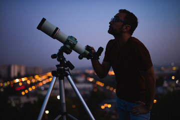 Astronomer with a telescope watching at the stars and Moon with blurred city lights in the...