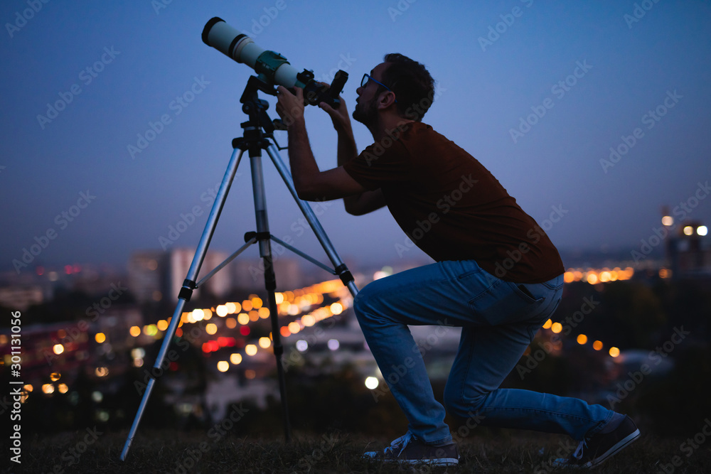 Wall mural astronomer with a telescope watching at the stars and moon with blurred city lights in the backgroun