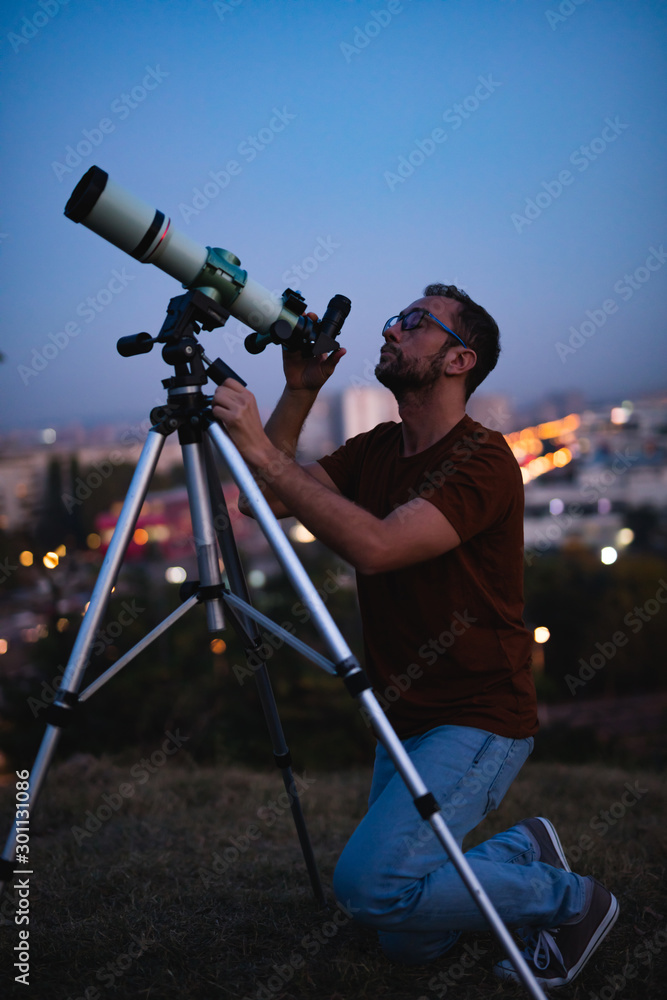 Wall mural astronomer with a telescope watching at the stars and moon with blurred city lights in the backgroun