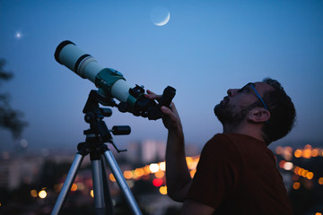 Astronomer with a telescope watching at the stars and Moon with blurred city lights in the...