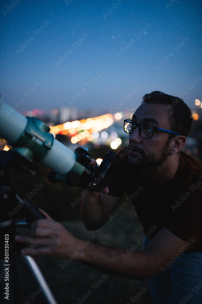 Wall mural astronomer with a telescope watching at the stars and moon with blurred city lights in the backgroun