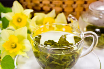 Green tea in a glass cup with tea leaves, yellow daffodil flowers and wicker basket on a white background.