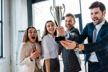 selective focus of excited multicultural businesswomen and businessmen looking at trophy in office - obrazy, fototapety, plakaty
