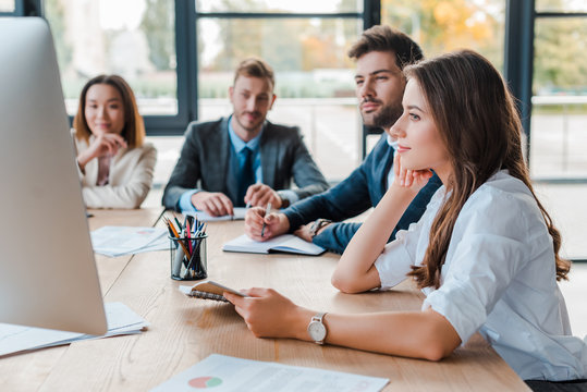selective focus of attractive businesswoman looking at computer during webinar monitor near multicultural coworkers in office