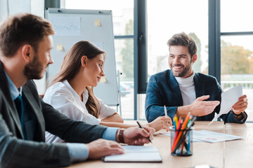 selective focus of cheerful businessman holding paper while sitting near coworkers in office