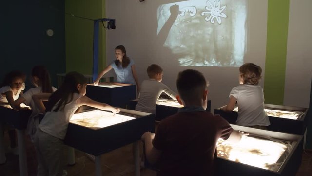 Young female art teacher showing how to make sand animation to group of children standing by lightboxes and working on their projects