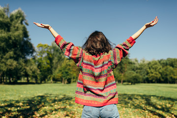 Rear view horizontal outdoor image of beautiful young woman windy hair enjoying the weather with wide arms open, wearing colorful knitted sweater, posing on nature sunlight background.