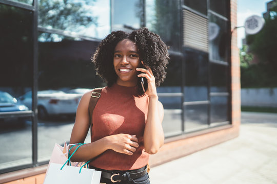 Portrait of carefree dark skinned female shopaholic with shopping bags in hand calling to friend for communicating about sales connected to roaming internet on cellphone, concept of Black Friday