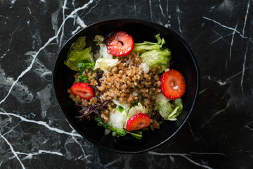 Buckwheat Salad with Strawberry Slices and Green Leaves in Black Bowl on Dark Granite Surface.
