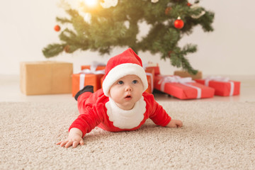 Obraz na płótnie Canvas Cute baby girl wearing santa claus suit crawling on floor over Christmas tree. Holiday season.
