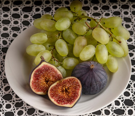 On a white plate are a brush of white grapes and fig fruits. Figs whole and cut in half. Close-up on a white patterned tablecloth.