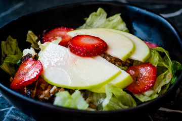 Apple Salad with Strawberry Slices, Buckwheat and Green Leaves in Black Bowl on Dark Granite Surface.