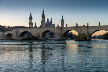 Fototapeta na wymiar View of the medieval stone bridge in front of the Cathedral of El Pilar, Zaragoza (Spain), on the banks of the River Ebro, during the sunset.