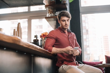 handsome man sitting near bar counter in wireless earphones using smartphone