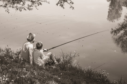 Back view of happy family on summer vacations concept. Father and son fishing together at river bank at scenic landscape background of fresh green grass and blue calm water. Horizontal sepia photo.