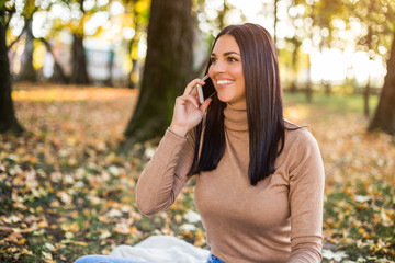 Beautiful woman talking on the phone while sitting in the park and enjoys in autumn.	