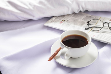 A cup of coffee and a newspaper on a white bed in the morning.