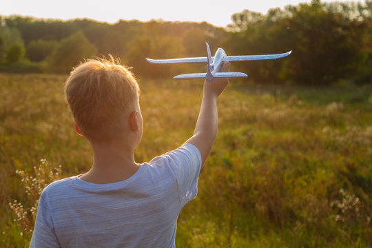 Back silhouette of cute happy cheerful child running fastly along grassy hill at countryside holding big toy plane in hand. Boy playing during sunset time in evening. Horizontal color photography.