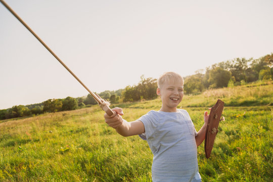 Closeup portrait of happy cheerful smiling white kid playing happily outdoor holding wooden sword and shield in hands. Horizontal color photography.