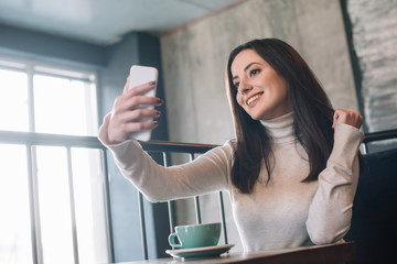 smiling young woman sitting at wooden table with cappuccino and taking selfie on smartphone on balcony in coffee shop