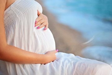 Young beautiful pregnant woman relax by the calm sunset sea on the sea shore touching her belly with love and care.