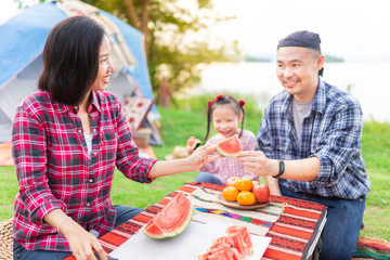 asian family picnic and camping in forest, asian female use knife slice watermelon and send to her husband, they relax and feeling happy, happiness family activity