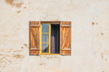 There is a window in the old plastered wall of the house. Stucco light color. One casement window and shutters open. The window is curtained. Background. Texture.