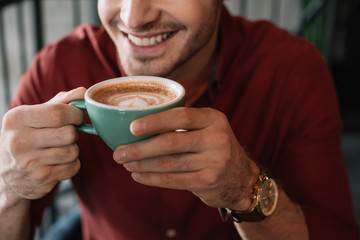 cropped view of smiling young man with cup of tasty cappuccino in coffee shop