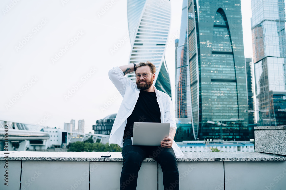 Poster portrait of bearded software developer sitting with modern laptop device and smiling at camera, happ