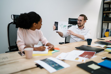 Positive man in eyeglasses sitting at office desk and showing paper with information during multicultural conference with afro american colleague, male and female partners enjoying collaboration