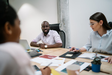 Successful diverse male and female colleagues cooperating near meeting table enjoying briefing togetherness, multicultural executive entrepreneurs dressed in formal wear discussing details of project