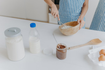 Cropped view of kid in apron whisks ingredients in bowl with beater, busy cooking and helping mother to prepare cake. Dough, milk, eggs, wooden spatula and chocolate on table