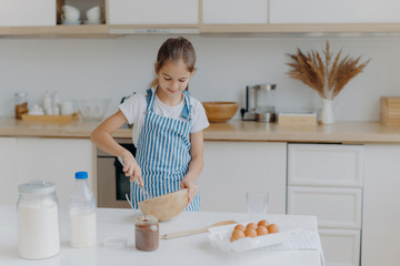 Cute small girl wears striped apron, whisks ingredients in bowl, prepares dough, teaches to cook, stands at white table with eggs, milk, flour against kitchen background. Childern and cooking concept