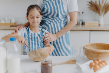 Cropped image of affectionate mother in apron embraces daughter who learns how to make pasty, holds whisk, mixes ingredients in bowl, poses at kitchen table with milk, melted chocolate, eggs