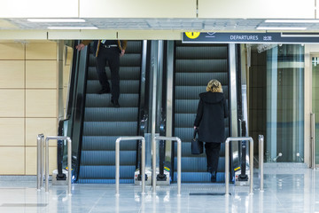 The alone woman on the escalator or moving staircase with inscription departure in English and Chinese in the international airport or railway station from the back moving upstairs with luggage