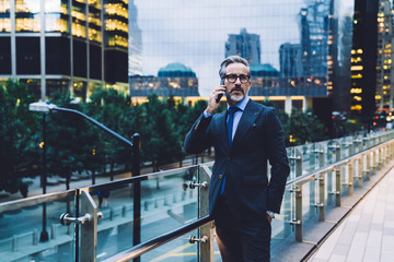 Adult businessman talking on phone near glass fence against evening New York street