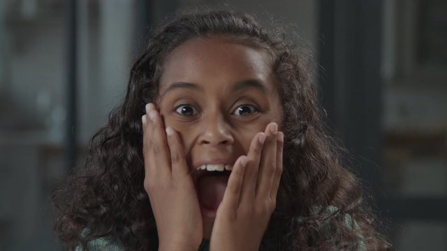 Portrait of emotionally stressed cute elementary age african girl with curly hair holding hands on cheeks, screaming loudly with mouth wide opened, expressing shock, fear and scare indoors.