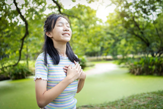 Happy smiling asian child girl standing in green nature,holding hands on heart,female teenage touching her chest,enjoy breathing fresh air with closed eyes,relaxing in park,healthy lifestyle concept