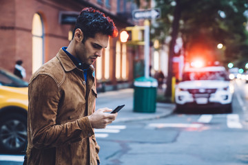 Thoughtful ethnic young man in jacket texting and walking along street