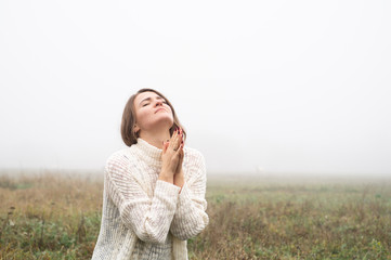 Girl closed her eyes on the knees, praying in a field during beautiful fog. 