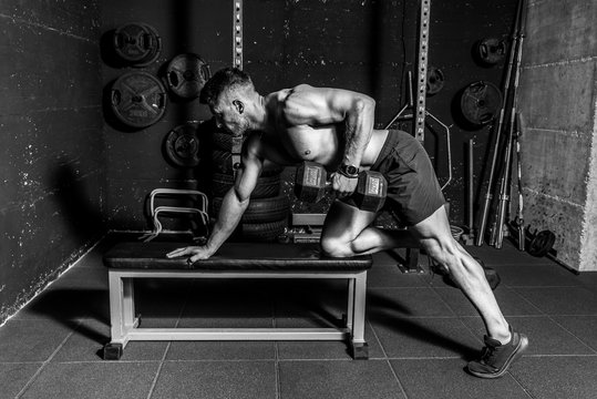 Young sweaty fit muscular strong man doing workout training back muscles with dumbbell on the bench in the gym dark image real people black and white