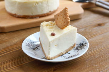 A piece of classic New York cheesecake decorated with square cookies on plate, on wooden table in coffee pastry shop with whole cake in the background. Cake sweet dessert for holiday, birthday party